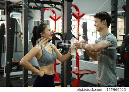 A woman and her trainer share a cheerful moment after a workout, clinking water bottles at the gym. A woman and her trainer share a cheerful moment after a workout, clinking water bottles at the gym. 135960913