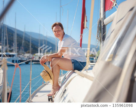 Mature woman relaxes on private yacht during sunny day in Finike Marina. Turkish coastline and luxury boats surround sailing vessel in Mediterranean port during summer vacation. 135961098