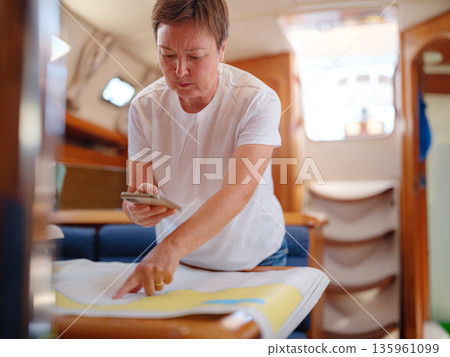 Mature woman sits inside yacht cabin analyzing paper sea chart for route planning. Navigation process involves calculating coordinates and path for upcoming sailing journey from Finike. 135961099