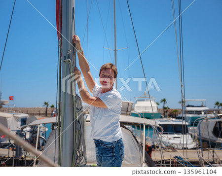 Female sailor holds white sail cloth preparing to hoist sails on yacht. Physical activity and boat handling skills are shown during preparation for sailing trip near Finike coast. Female sailor holds white sail cloth preparing to hoist sails on yacht. Physical activity and boat handling skills are shown during preparation for sailing trip near Finike coast. 135961104