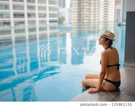 Young asian woman relaxing by pool at Kuala Lumpur hotel with view of surrounding skyscrapers, enjoying leisure time in vibrant urban setting. Young asian woman relaxing by pool at Kuala Lumpur hotel with view of surrounding skyscrapers, enjoying leisure time in vibrant urban setting. 135961110