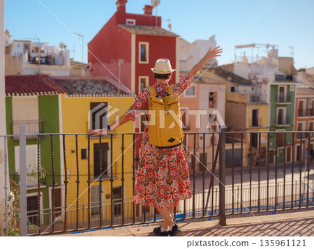 Woman in dress strolls through colorful streets of Spanish coastal town of La Vila Joiosa . sunny winter atmosphere highlights charm of Mediterranean architecture and quiet seaside life, back view 135961121