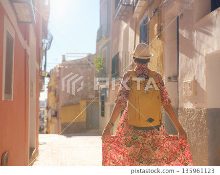 Woman in dress strolls through colorful streets of Spanish coastal town of La Vila Joiosa . sunny winter atmosphere highlights charm of Mediterranean architecture and quiet seaside life, back view Woman in dress strolls through colorful streets of Spanish coastal town of La Vila Joiosa . sunny winter atmosphere highlights charm of Mediterranean architecture and quiet seaside life, back view 135961123