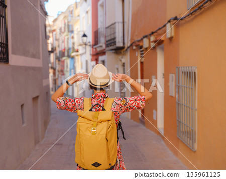 Woman in dress strolls through colorful streets of Spanish coastal town of La Vila Joiosa . sunny winter atmosphere highlights charm of Mediterranean architecture and quiet seaside life, back view 135961125