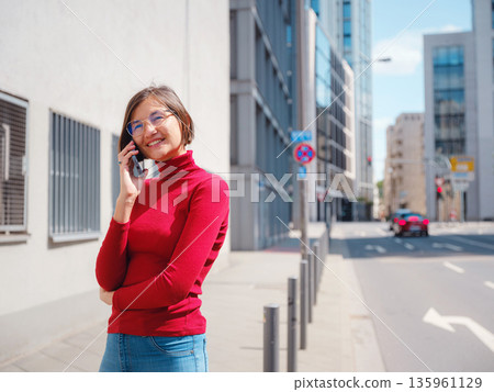 Woman in glasses and red turtleneck, speaks on phone in center of Frankfurt, surrounded by modern buildings and fast-paced city life, balancing professionalism and communication in urban setting. Woman in glasses and red turtleneck, speaks on phone in center of Frankfurt, surrounded by modern buildings and fast-paced city life, balancing professionalism and communication in urban setting. 135961129