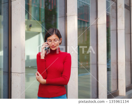Woman in glasses and red turtleneck, speaks on phone in center of Frankfurt, surrounded by modern buildings and fast-paced city life, balancing professionalism and communication in urban setting. Woman in glasses and red turtleneck, speaks on phone in center of Frankfurt, surrounded by modern buildings and fast-paced city life, balancing professionalism and communication in urban setting. 135961134