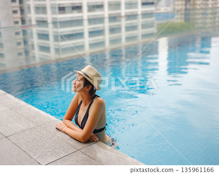 Young asian woman relaxing by pool at Kuala Lumpur hotel with view of surrounding skyscrapers, enjoying leisure time in vibrant urban setting. Young asian woman relaxing by pool at Kuala Lumpur hotel with view of surrounding skyscrapers, enjoying leisure time in vibrant urban setting. 135961166