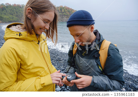 Couple enjoys Turkish coastline. walking in stone beach, watching sea waves, relaxing together, slow romantic moment near calm blue water under soft winter light. Couple enjoys Turkish coastline. walking in stone beach, watching sea waves, relaxing together, slow romantic moment near calm blue water under soft winter light. 135961183