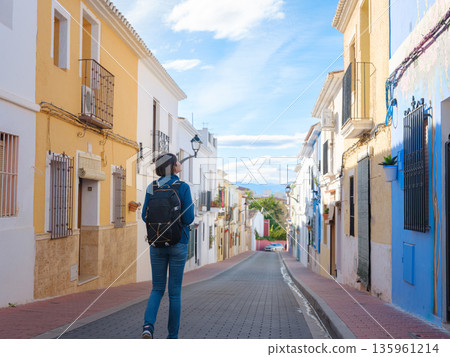 Young woman strolling through charming narrow streets of Denia, Alicante province, Spain, surrounded by historic Mediterranean architecture , enjoying peaceful atmosphere and cultural ambiance. Young woman strolling through charming narrow streets of Denia, Alicante province, Spain, surrounded by historic Mediterranean architecture , enjoying peaceful atmosphere and cultural ambiance. 135961214