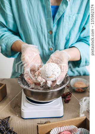 Hands hold round bath bomb over metal bowl on digital scale surrounded by packaging materials. Bath bomb making, handcrafted cosmetics, artisan soapery, small batch beauty, DIY bath products. Hands hold round bath bomb over metal bowl on digital scale surrounded by packaging materials. Bath bomb making, handcrafted cosmetics, artisan soapery, small batch beauty, DIY bath products. 135962374