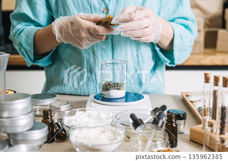 Person in protective gloves adds botanical ingredients to glass beaker on digital scale. Micro-manufacturing, artisan lab, small-scale cosmetics. 135962385