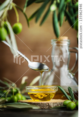 Pouring olive oil from spoon into glass bowl with olives and bottle on rustic table 135962446