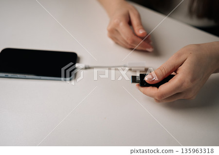 Closeup view of unrecognizable woman connecting usb flash drive to smartphone through adapter cable, facilitating data transfer and expanding mobile storage sitting on clean white table. 135963318