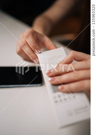 Vertical cropped shot of woman opening white retail box containing new phone cable adapter, holding over clean white table with smartphone positioned nearby, showcasing excitement and anticipation. 135963320