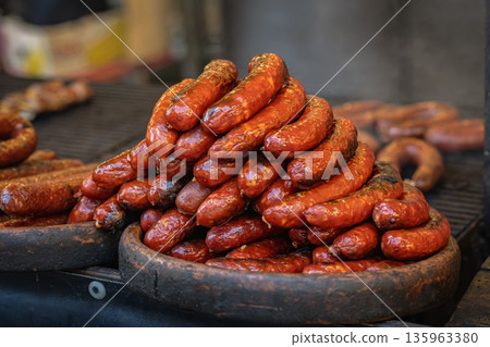 Pile of sausages at food stand. Grilled sausage on tray, barbecue grill with fire heat and smoke, street food market stall. Concept of sausage street food cooking, catering service, food marketing 135963380