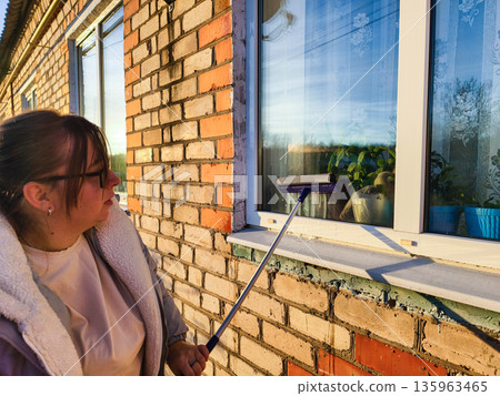 A woman doing housework carefully cleans a dirty window with a squeegee. The woman's cleaned window reflects the sky and surrounding scenery on a sunny day. The woman is cleaning the window. A woman doing housework carefully cleans a dirty window with a squeegee. The woman's cleaned window reflects the sky and surrounding scenery on a sunny day. The woman is cleaning the window. 135963465
