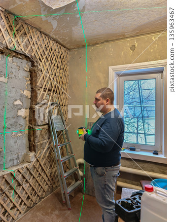 A man with a laser level projects green lines onto a wall with exposed wood siding and an unfinished ceiling while renovating an old room. A man is renovating a room in a house. 135963467