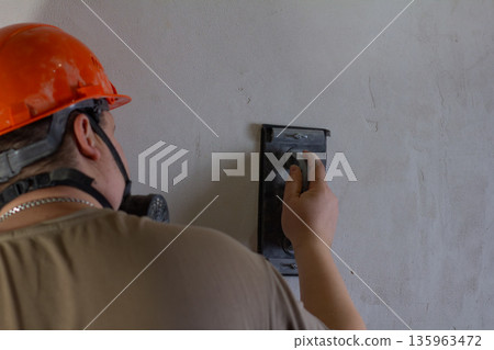 A construction worker wearing a safety helmet and a respirator mask carefully sands the rough surface of a wall with a manual grinder. A builder prepares a wall for painting during a house renovation. A construction worker wearing a safety helmet and a respirator mask carefully sands the rough surface of a wall with a manual grinder. A builder prepares a wall for painting during a house renovation. 135963472