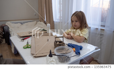 Child girl focused on DIY cardboard craft at table, using hot glue gun and scissors, doing hands-on learning and creative play at home. A child makes a craft from natural materials. Child girl focused on DIY cardboard craft at table, using hot glue gun and scissors, doing hands-on learning and creative play at home. A child makes a craft from natural materials. 135963475