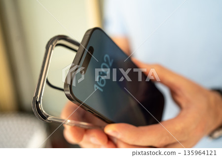 Close-up of a man holding a smartphone and a transparent protective case, preparing to put the case on the phone. 135964121