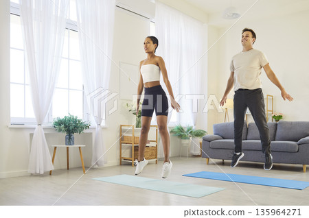 Smiling couple doing cardio jumps at home, staying active, healthy in living room Smiling couple doing cardio jumps at home, staying active, healthy in living room 135964271