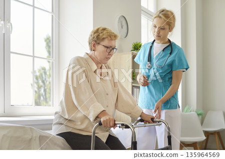 Senior woman and caregiver in medical room, young nurse assisting older patient sitting on bed 135964569