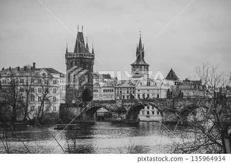Visitors walk along the Charles Bridge while admiring the Tower. Trees surround the area as the river reflects the historic buildings nearby during cloudy weather. Visitors walk along the Charles Bridge while admiring the Tower. Trees surround the area as the river reflects the historic buildings nearby during cloudy weather. 135964934