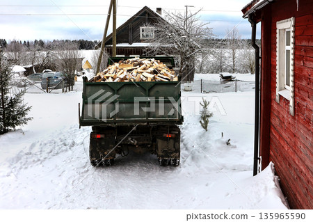 Dump truck prepares to unload split logs in front of rural house in snowy winter village. Dump truck prepares to unload split logs in front of rural house in snowy winter village. 135965590