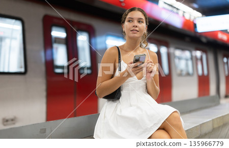 Positive young girl sitting on subway platform holding phone in hand Positive young girl sitting on subway platform holding phone in hand 135966779