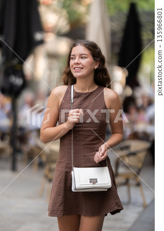 Cute young girl beautifully dressed in modern summer dress walking along city streets and choosing cafe to have snack and have good time Cute young girl beautifully dressed in modern summer dress walking along city streets and choosing cafe to have snack and have good time 135966801