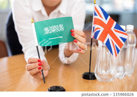 Female hands arranging flags of Saudi Arabia and United Kingdom on negotiating table Female hands arranging flags of Saudi Arabia and United Kingdom on negotiating table 135966826