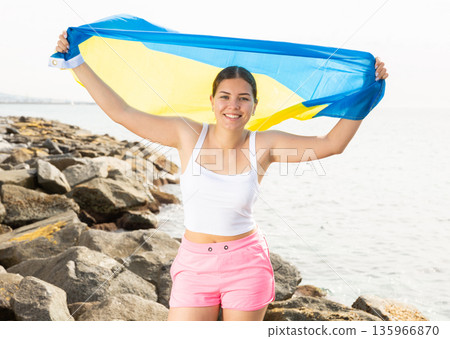 Young woman holding Ukraine waving flag at coastline against of bright sea Young woman holding Ukraine waving flag at coastline against of bright sea 135966870