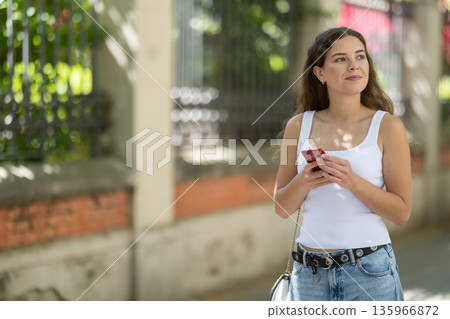 Happy woman looking to phone in hands and chatting in city 135966872