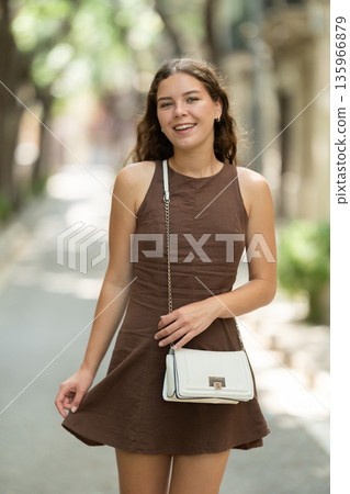 Cute young girl wearing stylish short summer dress with white handbag on shoulder walking along shady tree lined street enjoying spring day in Barcelona 135966879