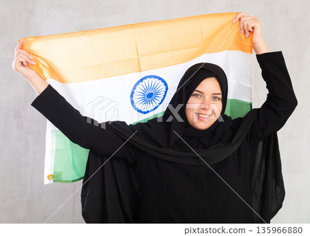 Balanced smiling Muslim woman in traditional black hijab holds flag of India 135966880