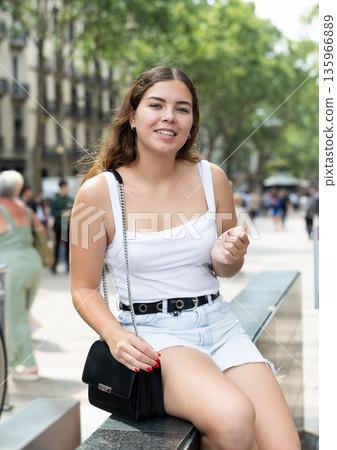 Happy young woman sitting on bench in Rambla Street of Barcelona 135966889