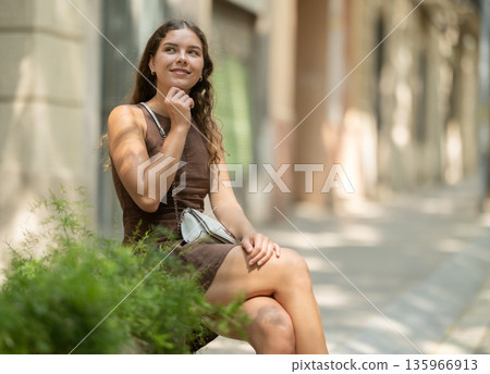 Young european girl sitting concrete flower bed with plants, relaxing and posing on shaded street in Barcelona 135966913