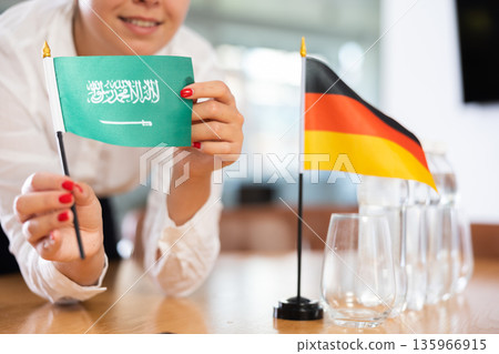 Little flag of Germany on table and flag of Saudi Arabia put next to it by young woman Little flag of Germany on table and flag of Saudi Arabia put next to it by young woman 135966915