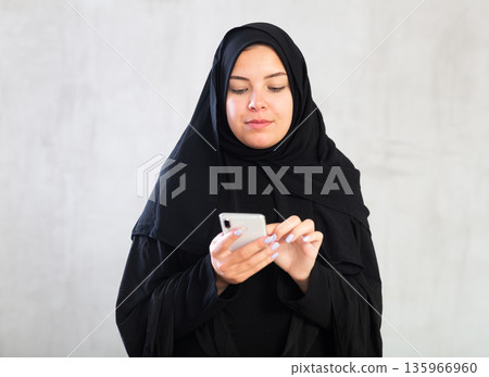 young positive girl in black hijab holds phone in her hands and surf Internet young positive girl in black hijab holds phone in her hands and surf Internet 135966960