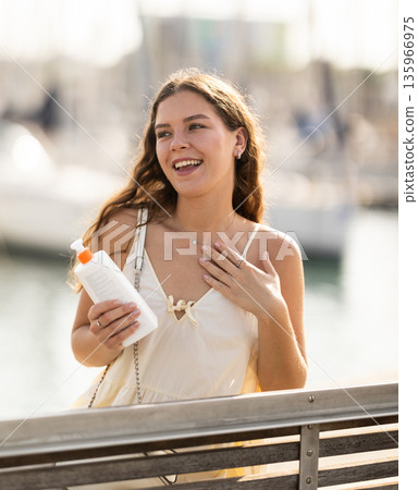Young brunette girl applying sunscreen to her skin while walking in city Young brunette girl applying sunscreen to her skin while walking in city 135966975