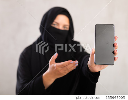 Young woman dressed in traditional islamic clothes with her face covered showing new phone model 135967005