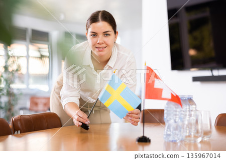 Positive young woman putting flag of Sweden on the table with flag of Switzerland in conference room Positive young woman putting flag of Sweden on the table with flag of Switzerland in conference room 135967014
