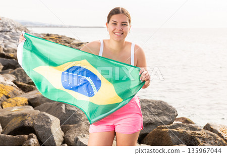 Pretty smiling girl holding Brazilian flag on the seashore on a sunny day Pretty smiling girl holding Brazilian flag on the seashore on a sunny day 135967044