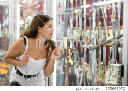Enthusiastic young girl looking at handicraft swords in souvenir shop Enthusiastic young girl looking at handicraft swords in souvenir shop 135967052