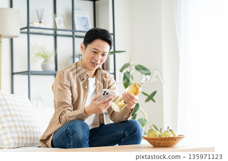 Middle-aged man holding smartphone and wine bottle in living room 135971123