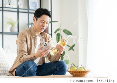Middle-aged man holding smartphone and wine bottle in living room 135971124