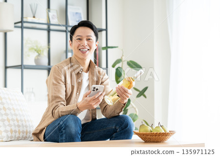 Middle-aged man holding smartphone and wine bottle in living room Middle-aged man holding smartphone and wine bottle in living room 135971125