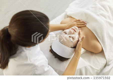 Young woman in spa salon receiving beauty procedure with moisturizing mask from beautician 135971502
