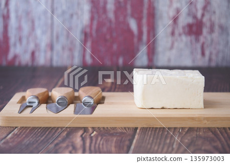 Cheese with knives on wooden board in kitchen setting 135973003