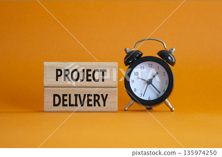 Project Delivery symbol. Concept word Project Delivery on wooden blocks. Beautiful orange background with alarm clock. Business and Project Delivery concept. Copy space 135974250